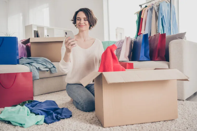 full size photo of cheerful young lady sit floor look phone donate clothes boxes apartment smile indoors in living room – mobile marketing reads