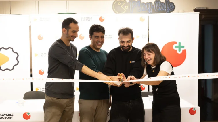 Four people cut a celebratory ribbon together, smiling, at an event backdrop with logos and a Playable Factory sign in the background.